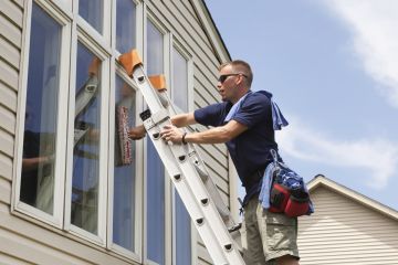Man on ladder cleaning exterior windows of a house under a blue sky